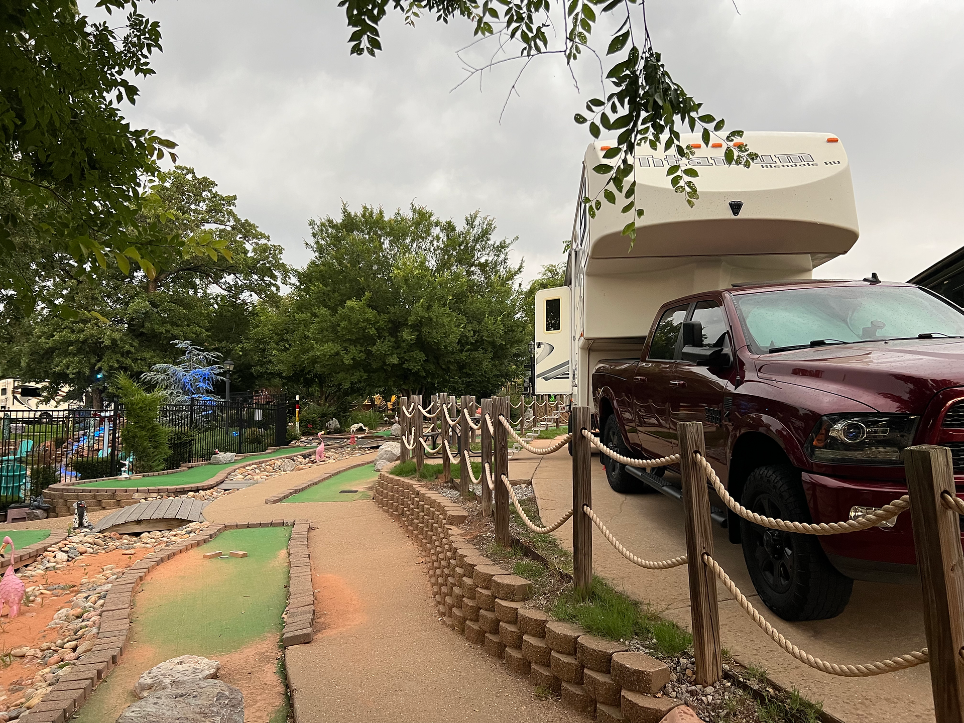 A charming campground scene featuring a red pickup truck hitched to a fifth-wheel RV trailer. The site includes a well-maintained mini-golf course with pathways bordered by rope fences, small bridges, and decorative elements like pink flamingos. Lush trees and greenery surround the area, creating a cozy and inviting atmosphere. The sky is overcast, indicating a cloudy day, which adds a serene and peaceful ambiance to the setting.