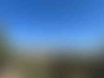 Expansive view of desert mountains and cliffs near Escalante, Utah, framed by juniper and pine trees in the foreground. The sunlit sandstone ridges stretch across the horizon beneath a vast, cloud-dotted blue sky.
