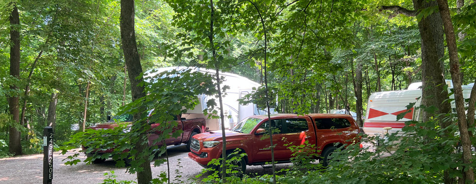 The image shows a well-shaded campsite in a dense, leafy forest. Two trucks are visible: a dark red pickup truck attached to a large white fifth-wheel trailer, and an orange pickup truck parked next to a white and red travel trailer. The campsite is surrounded by tall trees and lush green foliage, creating a tranquil and secluded atmosphere. A campsite marker with the numbers "249" and "250E" is seen in the foreground. The gravel ground and dappled sunlight filtering through the trees add to the serene and inviting nature of the campsite.