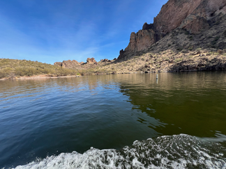 View from a moving boat on Canyon Lake with rippling water in the foreground, rugged desert cliffs and hills in the background, and a bright blue sky overhead.