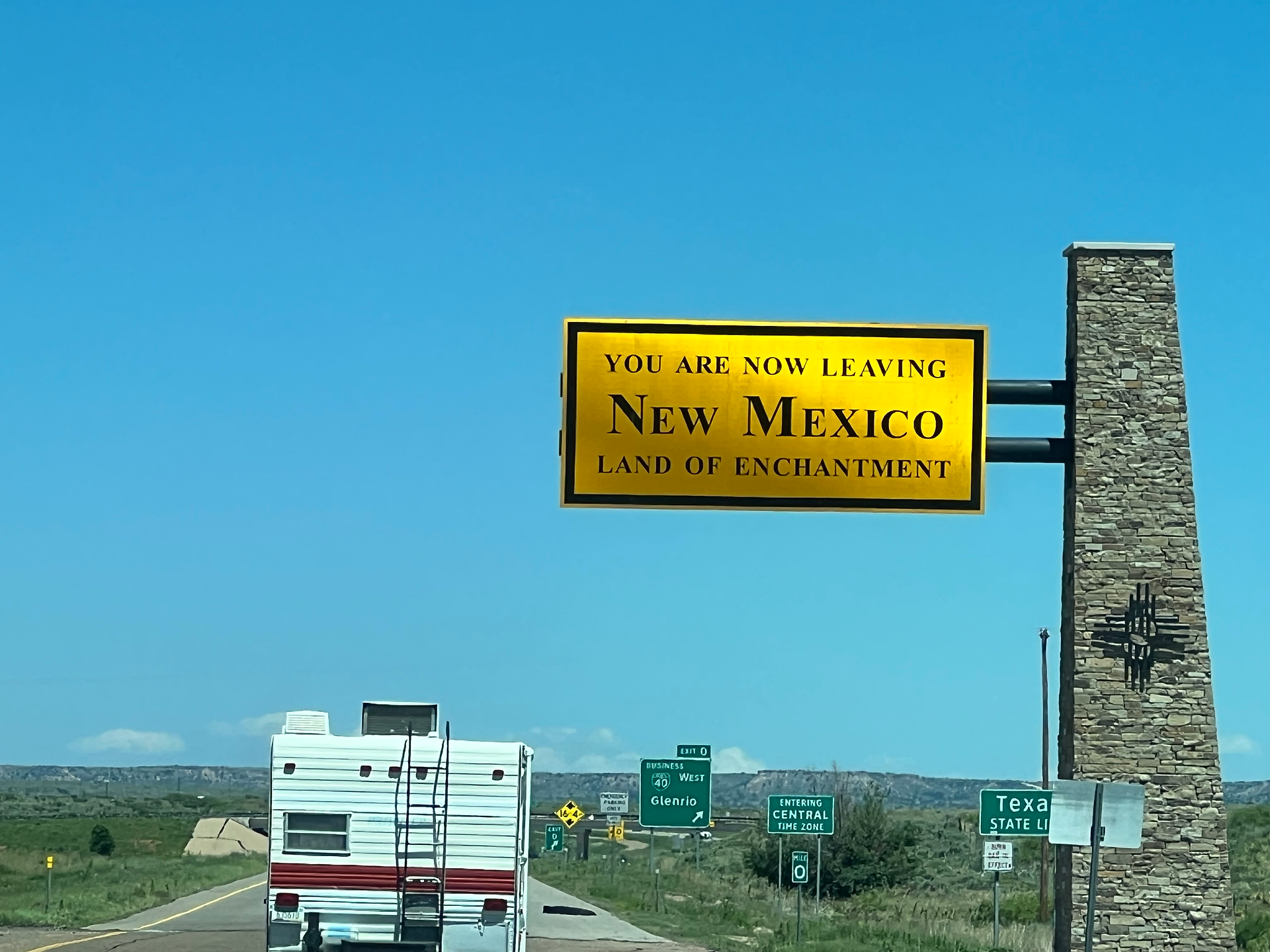 The image shows a highway scene where a large, yellow road sign reads "YOU ARE NOW LEAVING NEW MEXICO LAND OF ENCHANTMENT." The sign is supported by a stone pillar on the right side. Below the sign, a white and red RV is driving on the highway. To the right, there are smaller green signs indicating "Texas State Line," "Entering Central Time Zone," and "Business Loop I-40 Glenrio." The sky is clear and blue, and the landscape in the background is open with low vegetation and distant hills.
