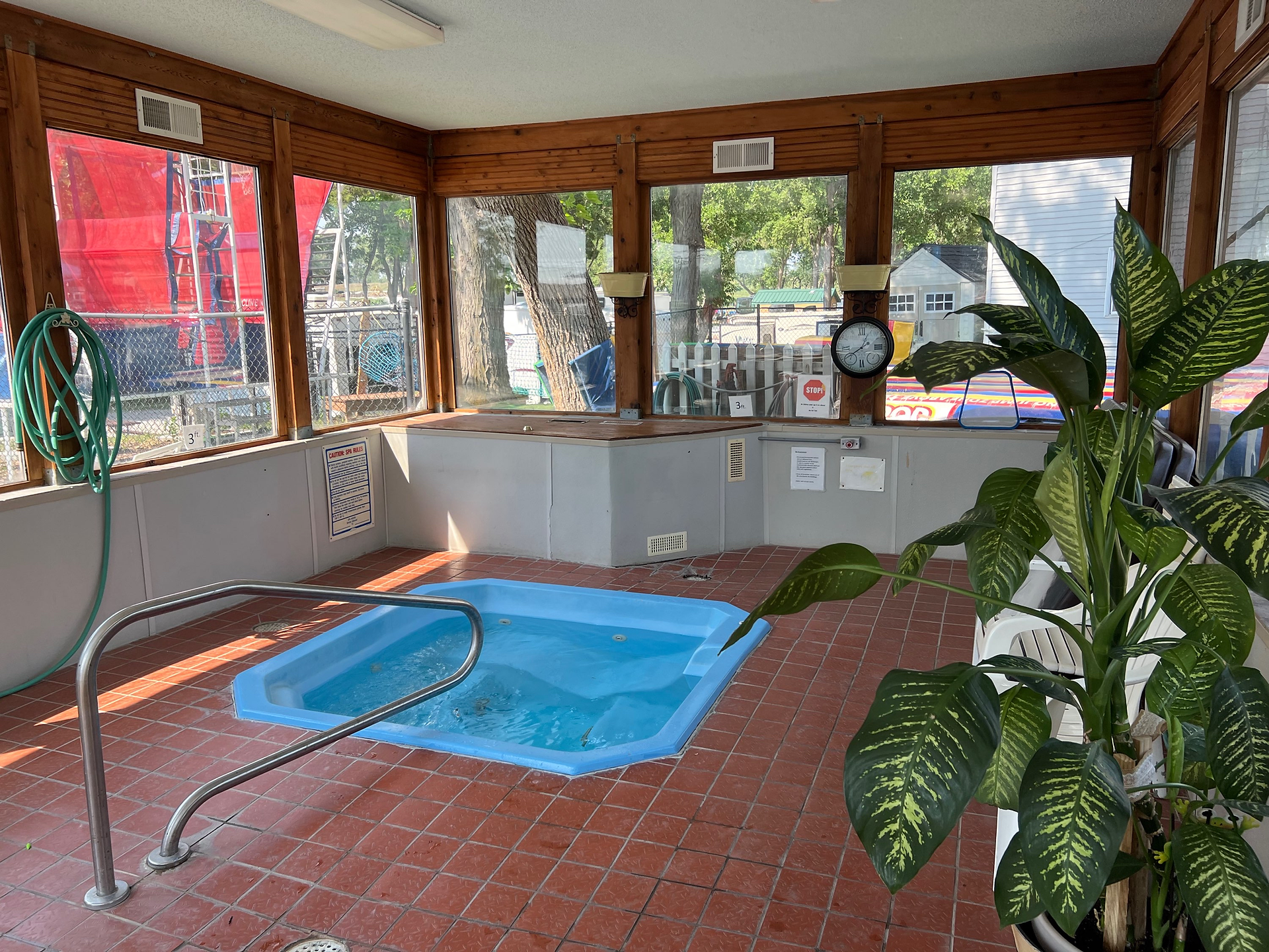 This image shows an indoor hot tub area within a glass-enclosed room. Key features include:  Hot Tub: The hot tub is a small, octagonal pool with blue water. It is set into a red-tiled floor and has metal handrails for safe entry and exit.  Glass Enclosure: The room is enclosed with large glass windows, allowing plenty of natural light to enter and providing a view of the outdoor area.  Plants: There are large green plants placed around the room, adding to the aesthetic appeal and creating a more inviting atmosphere.  Wall Fixtures: A hose is coiled on the wall, suggesting it is used for cleaning or maintenance. There are also several signs and notices on the walls, possibly indicating rules or safety instructions.  Clock: A clock is mounted on the wall, helping users keep track of time while enjoying the hot tub.  Outdoor View: Through the windows, you can see a chain-link fence and some outdoor structures, indicating that this hot tub area is part of a larger recreational facility.  The overall setting suggests a cozy and well-maintained indoor hot tub area, providing a relaxing environment for guests, likely part of a campground, RV park, or a similar recreational facility.