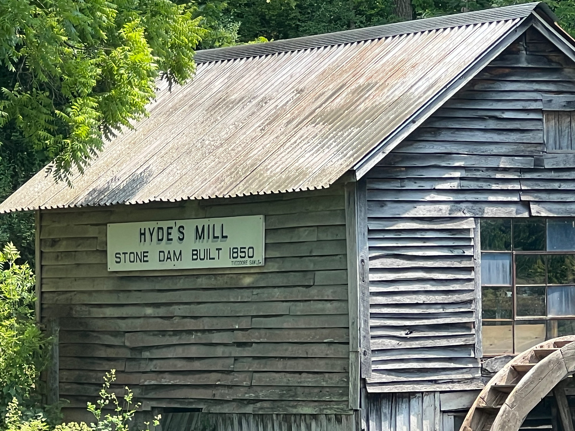 The image is a close-up view of the historic wooden mill known as "Hyde's Mill." The mill has a weathered, rustic appearance with aged wooden planks and a corrugated metal roof. A sign on the side of the building reads, "Hyde's Mill - Stone Dam Built 1850." The text below indicates "Tiogaore Sons."  The mill exudes a sense of history and timelessness, with its worn exterior and simple, sturdy construction. The window on the side of the mill reflects the surrounding greenery, adding to the natural and serene atmosphere. The lush green foliage surrounding the mill further enhances the picturesque and tranquil setting of this historic site.