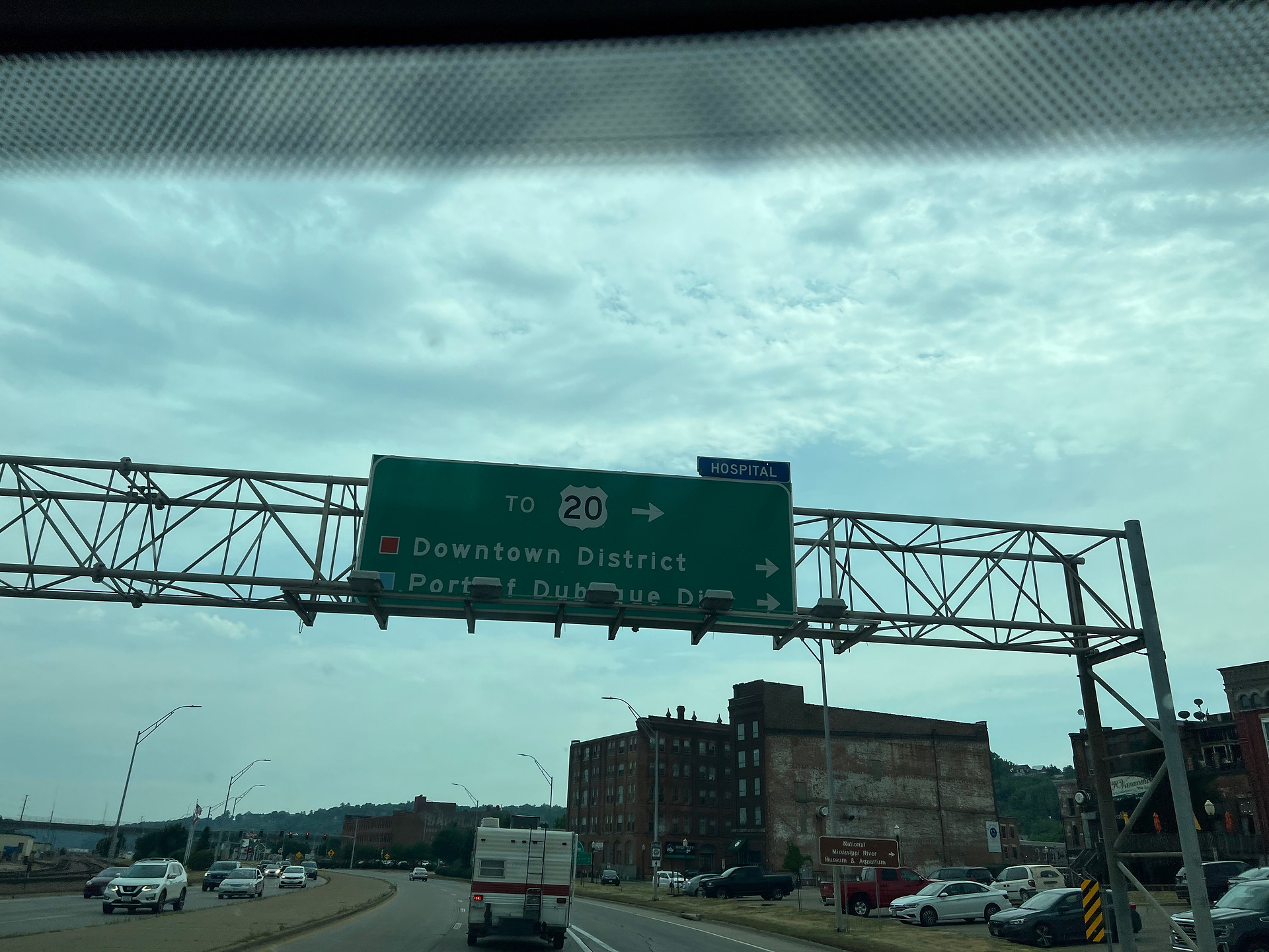 This image shows a highway sign directing drivers towards the Downtown District and the Port of Dubuque, indicating this area is a key part of the city. The sign also points towards the hospital, providing important directions for medical services.  The highway is busy with traffic, including a large RV in the foreground, suggesting the area may be frequented by travelers and tourists. In the background, the skyline features several old brick buildings, indicating an industrial or historical section of the city.  The overcast sky adds a neutral tone to the scene, making the green highway sign and the urban environment more prominent. This snapshot captures a moment of urban transit, highlighting the connections to vital parts of the city such as the downtown area and port.