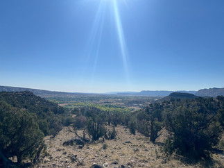 A sunbeam shines down over a vast desert valley near Escalante, Utah, illuminating a landscape of rolling hills, green trees, and distant mesas under a crystal-clear blue sky. The rugged terrain in the foreground is dotted with juniper trees and rocks.