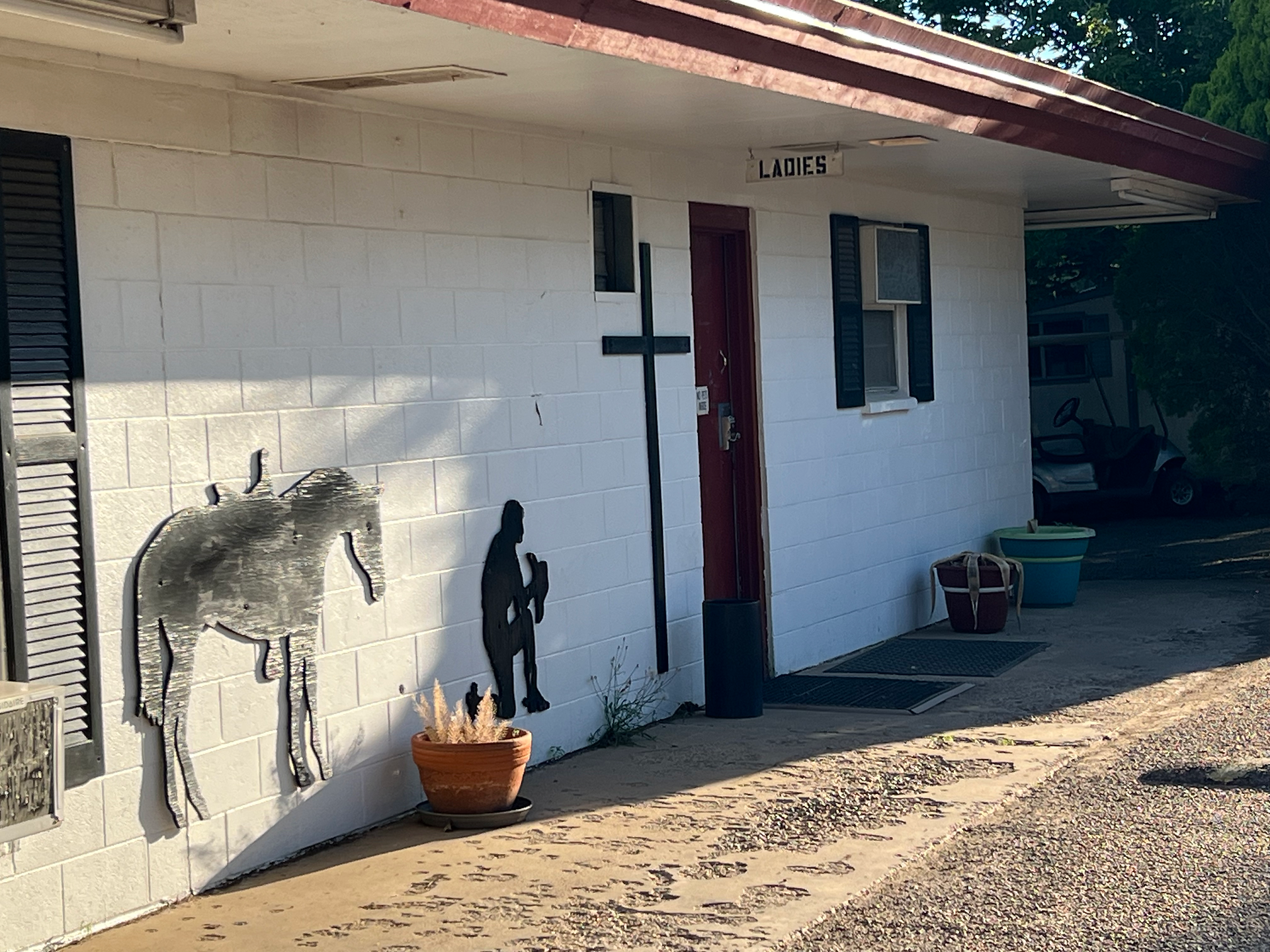 The exterior of a building labeled "LADIES," indicating it is a women's restroom or shower facility. The building has a white brick wall with a red door. The wall features black metal silhouettes of a horse and a person, adding a decorative touch. The area in front of the building is paved and includes some potted plants, contributing to a welcoming and rustic atmosphere. The building appears well-maintained and clean.