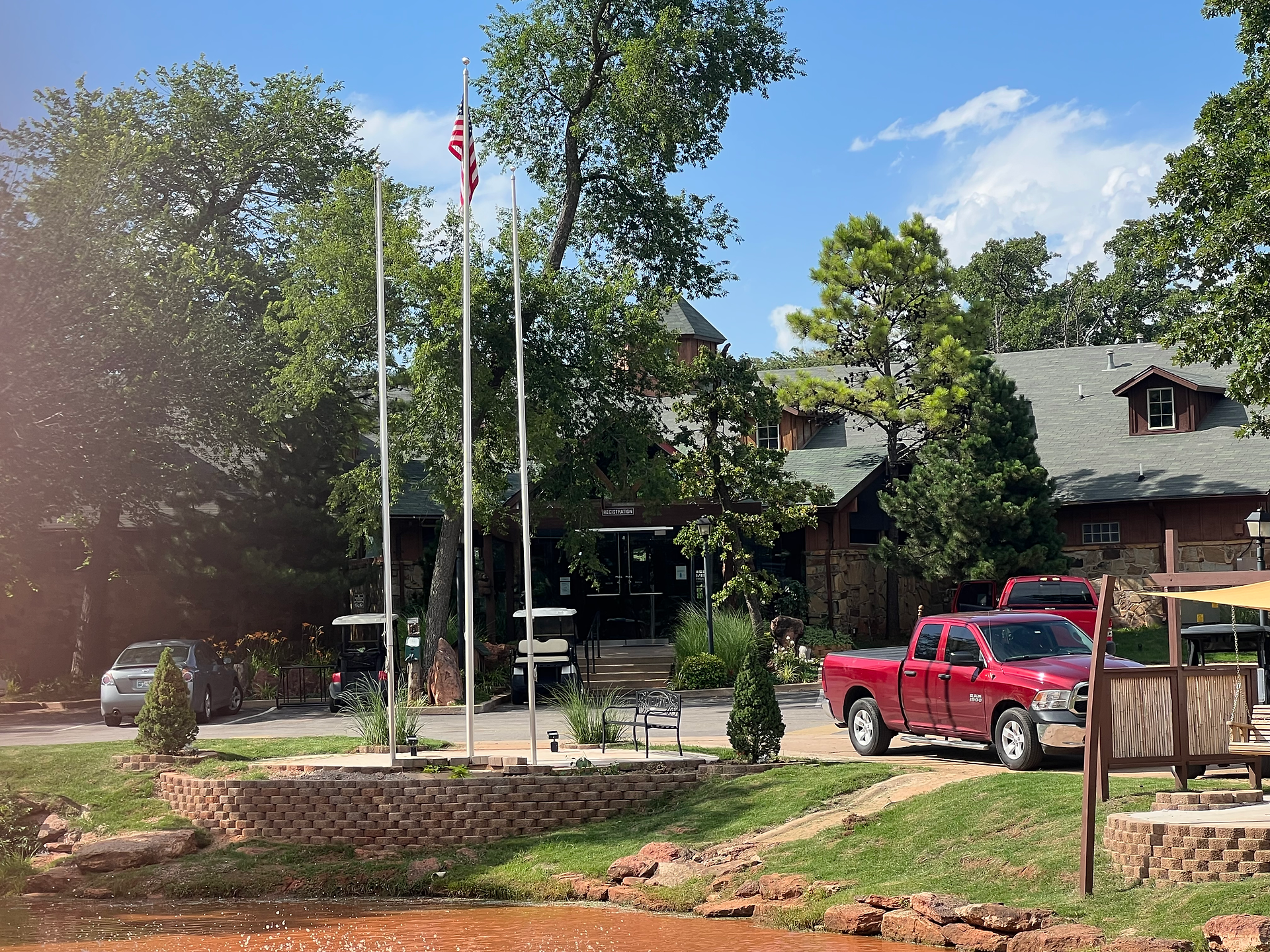 A picturesque view of the entrance to a campground, featuring the main building partially obscured by tall, lush trees. Three flagpoles are positioned at the front, with an American flag waving in the breeze. A red pickup truck is parked nearby, adding a pop of color to the serene setting. The landscaping includes neatly trimmed grass, a small brick retaining wall, and various plants, enhancing the welcoming and well-maintained appearance of the area. The clear blue sky and bright sunlight contribute to the inviting atmosphere.