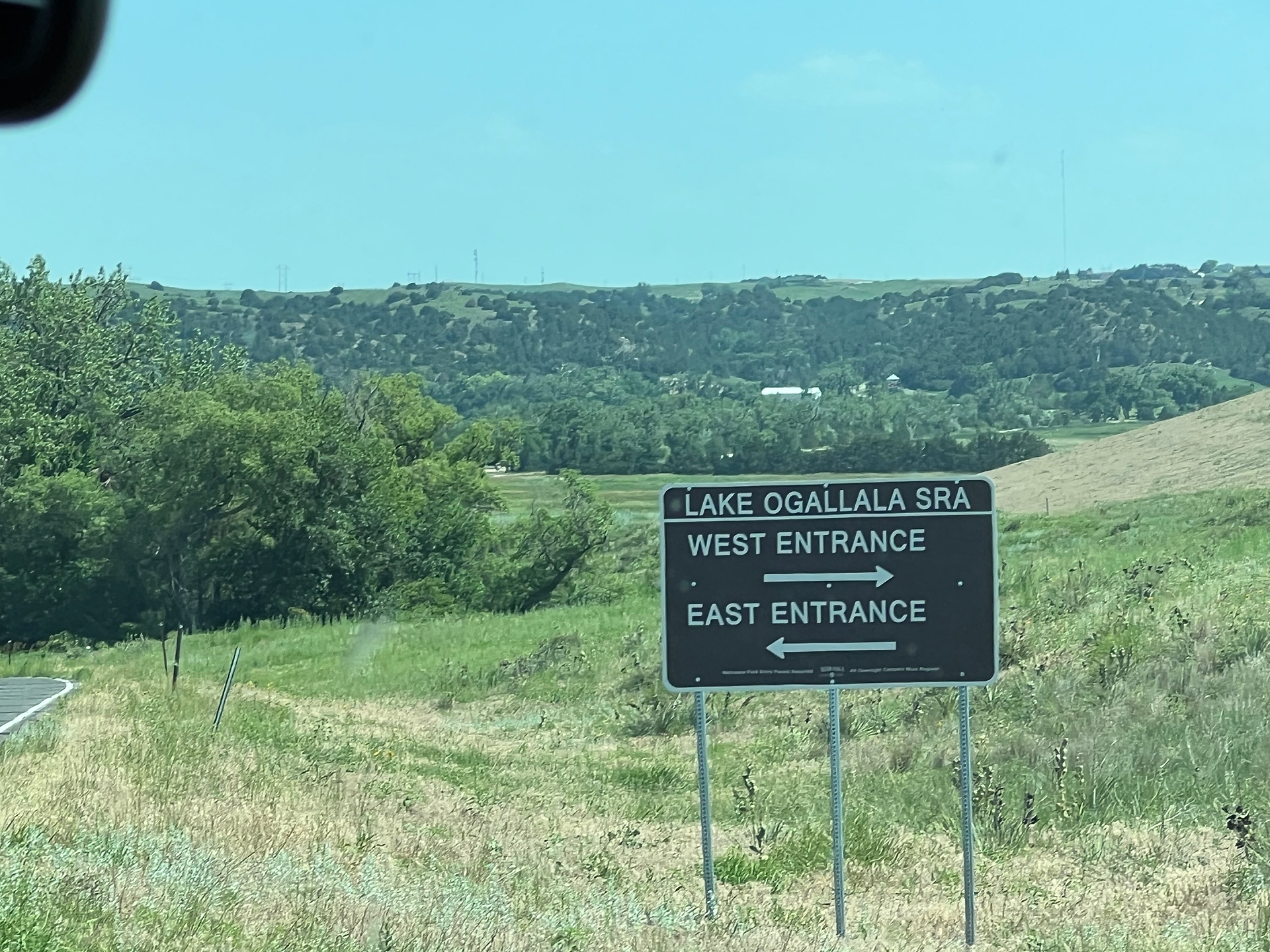 A scenic view of Lake Ogallala State Recreation Area with a directional sign in the foreground. The sign indicates the West Entrance to the right and the East Entrance to the left. Lush green hills and trees are visible in the background under a clear blue sky.