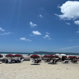 Sunny beach scene with rows of red-and-white umbrellas and lounge chairs facing the ocean, with gentle waves and a distant coastline under a bright blue sky.