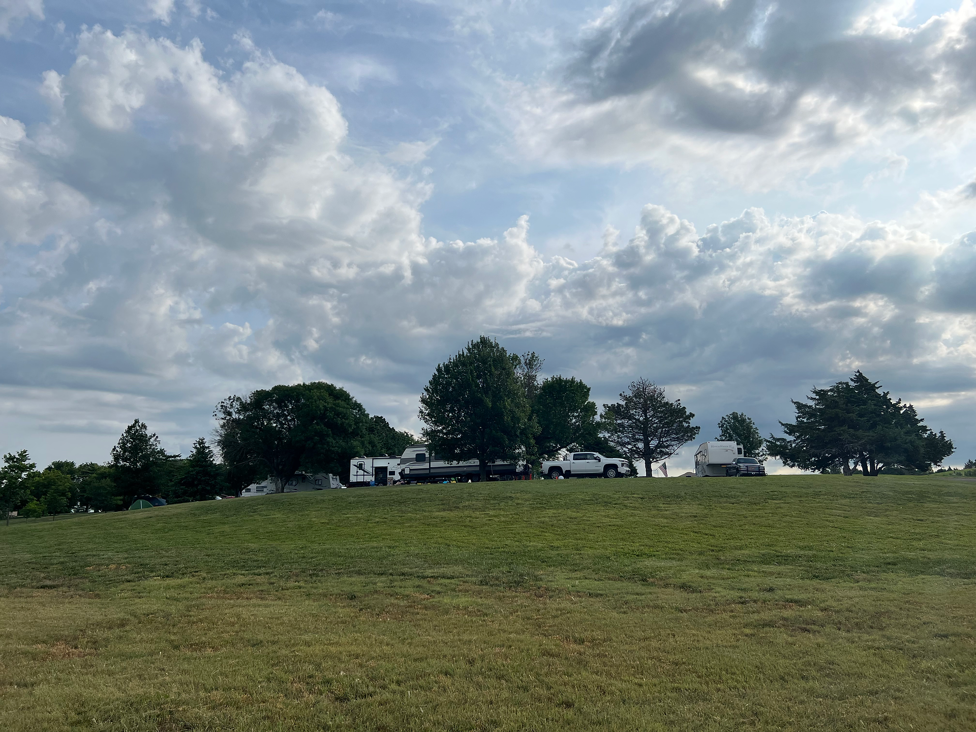A scenic view of a grassy hill with several trees at the top, under a partly cloudy sky. There are RVs and trucks parked along the ridge, indicating a campsite. The setting conveys a tranquil, open environment with a mix of clear skies and dramatic clouds above.