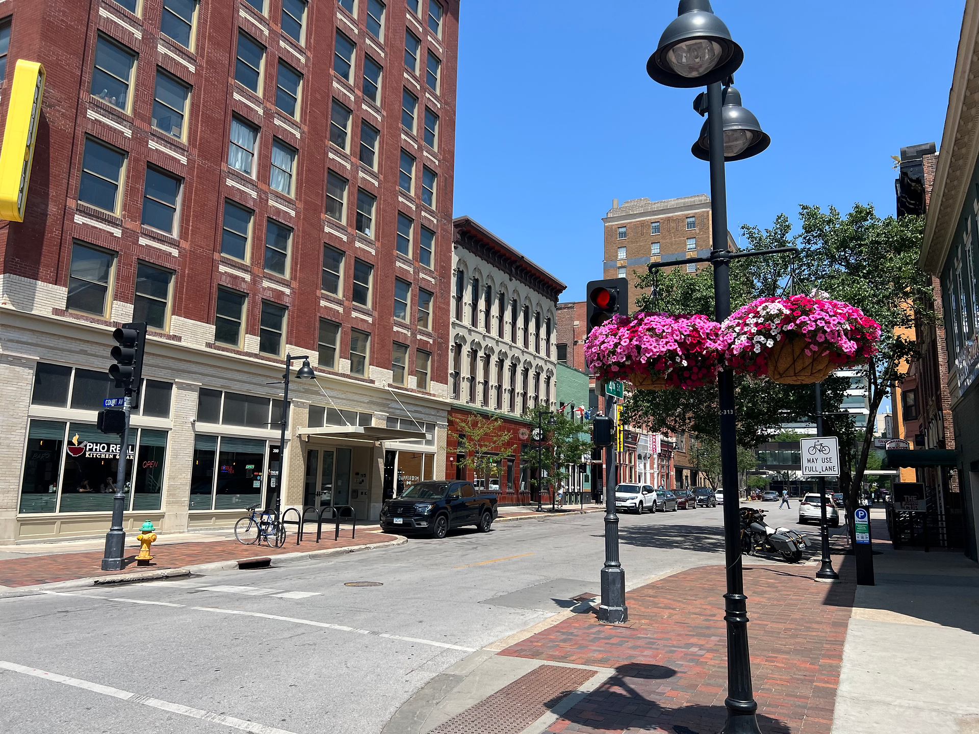 A vibrant city street scene featuring a row of historic buildings with various businesses, including a restaurant named "Pho Real Kitchen & Bar." The street is adorned with hanging baskets of bright pink flowers attached to black lampposts. The red-brick building facades and wide windows add charm to the area, while a bicycle is parked near a yellow fire hydrant. The clear blue sky and sunny weather enhance the lively and inviting atmosphere of the downtown setting.