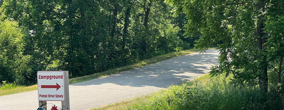 The image shows a small sign by the side of a paved road in a wooded area. The sign reads "Campground" with an arrow pointing to the right, and "Please Drive Slowly" below the arrow. Below the text, there is a decorative circular frame with colorful flowers and the word "WELCOME" at the bottom. The surrounding area is lush with greenery, tall trees, and a well-maintained grassy verge. The road curves gently to the right, leading further into the forested area, creating a serene and inviting atmosphere for campers.