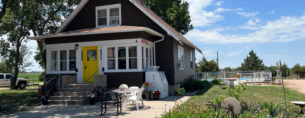 The image features a quaint and charming office building, likely part of a campground or RV park. The building is a cozy house with dark brown siding and a bright yellow front door that stands out, giving it a welcoming and friendly appearance. The sign indicating "OFFICE" is clearly visible, suggesting that this is where guests check in or seek assistance.  In front of the office, there is a concrete patio area with seating arrangements, including a white table and chairs, as well as some black chairs, inviting visitors to sit and relax. Several potted flowers and plants add to the homely and inviting atmosphere.  The surrounding area is well-maintained, with lush green grass and a variety of plants and flowers lining the walkway. There is also a large ice machine located near the entrance, which is a common feature at campgrounds and RV parks for guests' convenience.  To the right of the office, a fenced pool area can be seen, suggesting recreational facilities available for guests. The blue sky with scattered clouds and the overall sunny weather enhance the inviting and pleasant feel of the place.  In the background, there are some trees and a parked vehicle, indicating that this location might offer ample space for parking and outdoor activities, making it an ideal spot for a relaxing stay.