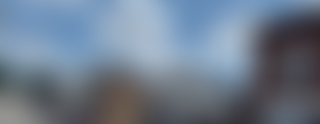 The image shows a street view of a small town with a variety of buildings lining the road. On the right, there is a brick building with a large window and an air conditioning unit in the upper window. Next to it is a white building with dark trim and a sloped roof. Further down the street, there are several other buildings, including a two-story building with a sign that appears to be a restaurant or cafe. The road is lined with parked cars, and a water tower can be seen in the background on the left side. The sky is clear with scattered clouds, creating a bright and sunny atmosphere. The overall scene depicts a quiet, small-town street with a mix of commercial and residential buildings.