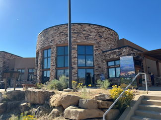The Escalante Visitor Center in Utah, a circular stone building with large windows and a modern rustic design, stands under a clear blue sky. A sign near the entrance welcomes visitors to the Grand Staircase–Escalante National Monument. Yellow wildflowers and desert plants grow among rocks in the foreground.