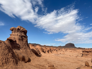 Sweeping desert view at Goblin Valley State Park in Utah, featuring layered red sandstone formations in the foreground and a flat-topped mesa in the distance beneath a vivid blue sky with scattered white clouds.