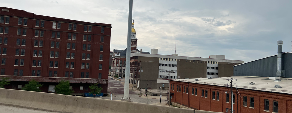 This image depicts an urban scene with several prominent buildings. In the foreground, there is a concrete barrier, likely part of a highway or overpass. Behind the barrier, several multi-story brick buildings can be seen. The building on the left appears to be an older industrial or warehouse structure with numerous windows.  In the center background, there's a historic building with a notable golden dome and a clock tower, adding a distinctive architectural feature to the scene. To the right, more modern buildings with flat roofs and fewer windows contrast with the older brick structures.  The sky remains overcast, which gives the scene a subdued, muted atmosphere. The overall composition captures a blend of historic and modern urban elements, reflecting the layered architectural history of the city.