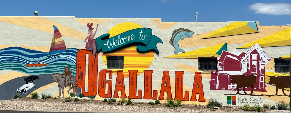 A colorful mural on a building wall welcoming visitors to Ogallala. The mural features various images, including a sailboat on blue waves, a cowboy on horseback, a jumping fish, and historic buildings. The text "Welcome to Ogallala" is prominently displayed, with the words "Keith County Chamber of Commerce" at the bottom. The vibrant artwork captures the spirit and attractions of the town under a clear blue sky.