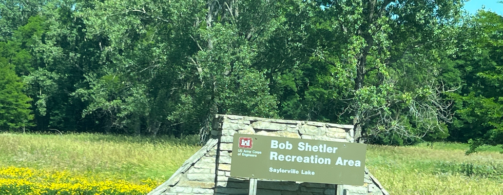 A sign for the Bob Shetler Recreation Area at Saylorville Lake, set against a backdrop of lush greenery and tall trees. The sign is mounted on a stone structure, with yellow wildflowers blooming at its base, adding a touch of color to the natural landscape. The bright, sunny day enhances the inviting atmosphere of the recreation area.