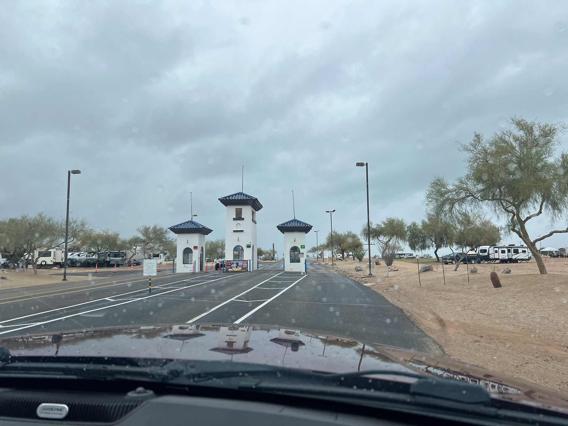 A view from inside a car approaching a toll gate entrance with three white booths topped with blue roofs. The area is surrounded by desert vegetation, RVs, and other vehicles. The sky is overcast, and raindrops are visible on the car windshield.