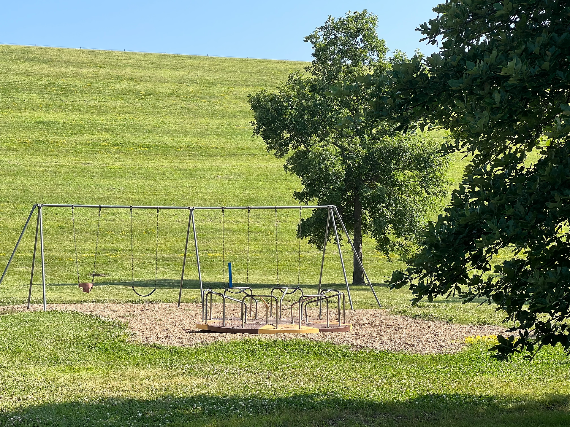 A quiet playground featuring a swing set and a merry-go-round, set on a grassy field. The area is surrounded by lush green grass and a few trees, with a large, gently sloping hill in the background. The playground equipment is positioned on a bed of wood chips, providing a safe play area for children. The clear blue sky and bright sunlight create a cheerful and inviting atmosphere.