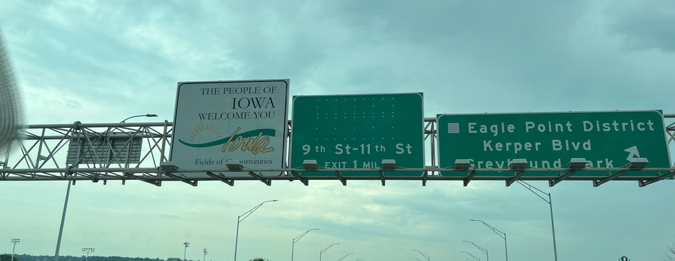 This image captures a highway scene with overhead signs indicating directions and welcoming travelers to Iowa. The signs include:  A welcome sign that reads, "The People of Iowa Welcome You" with the state name "Iowa" and the tagline "Fields of Opportunities." Directional signs for 9th St-11th St (Exit 1 Mile) and for the Eagle Point District, Kerper Blvd, and Greyhound Park. The sky is overcast, and the highway is relatively clear with a few vehicles visible, including a truck and a red car. The surrounding landscape is green with a few trees and buildings in the distance. The overall atmosphere suggests a typical highway entry point into a state, emphasizing a sense of travel and transition.