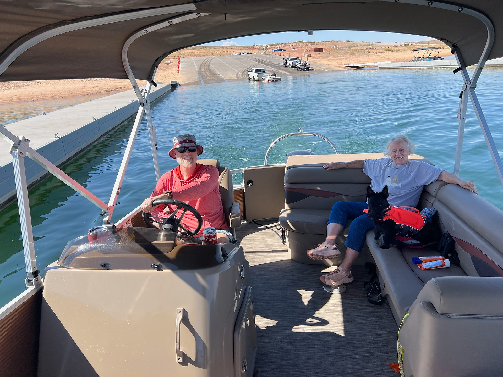 An elderly couple smiles while relaxing on a pontoon boat near a dock, with the man at the wheel wearing a red shirt and bucket hat, and the woman sitting on the bench beside a small black dog in an orange life vest. The sunny day and calm blue water create a cheerful boating scene.
