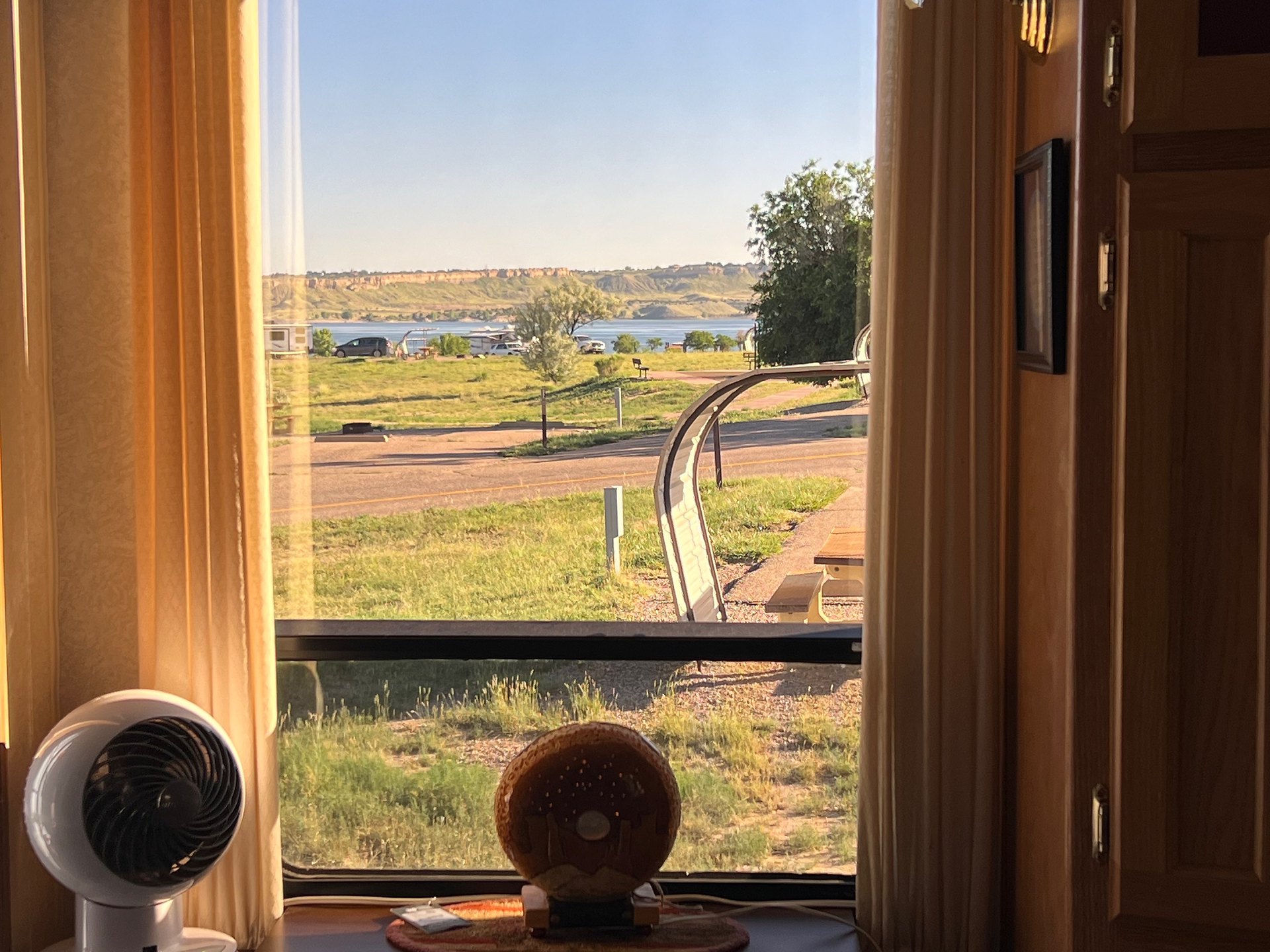 View of Lake Pueblo through a window with curtains framing the scene. Outside, there is a picnic table and grassy area, with the lake and hills visible in the distance under a clear sky. Inside, a small fan and a decorative item are placed on the windowsill, adding a cozy touch to the interior.
