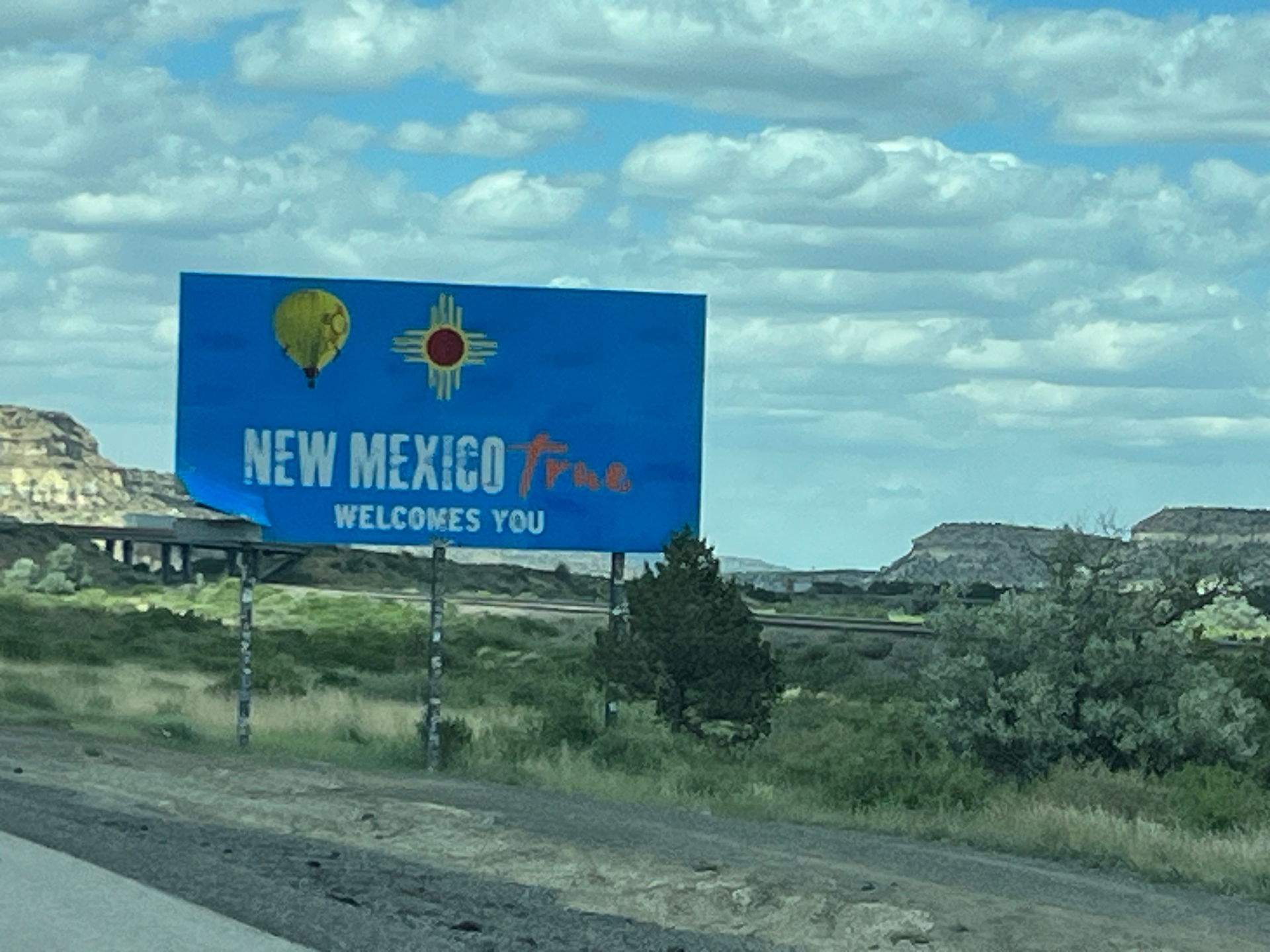 A blue road sign with 'New Mexico True Welcomes You' written on it, featuring a hot air balloon and the Zia sun symbol. The background shows a desert landscape with hills and a partly cloudy sky.