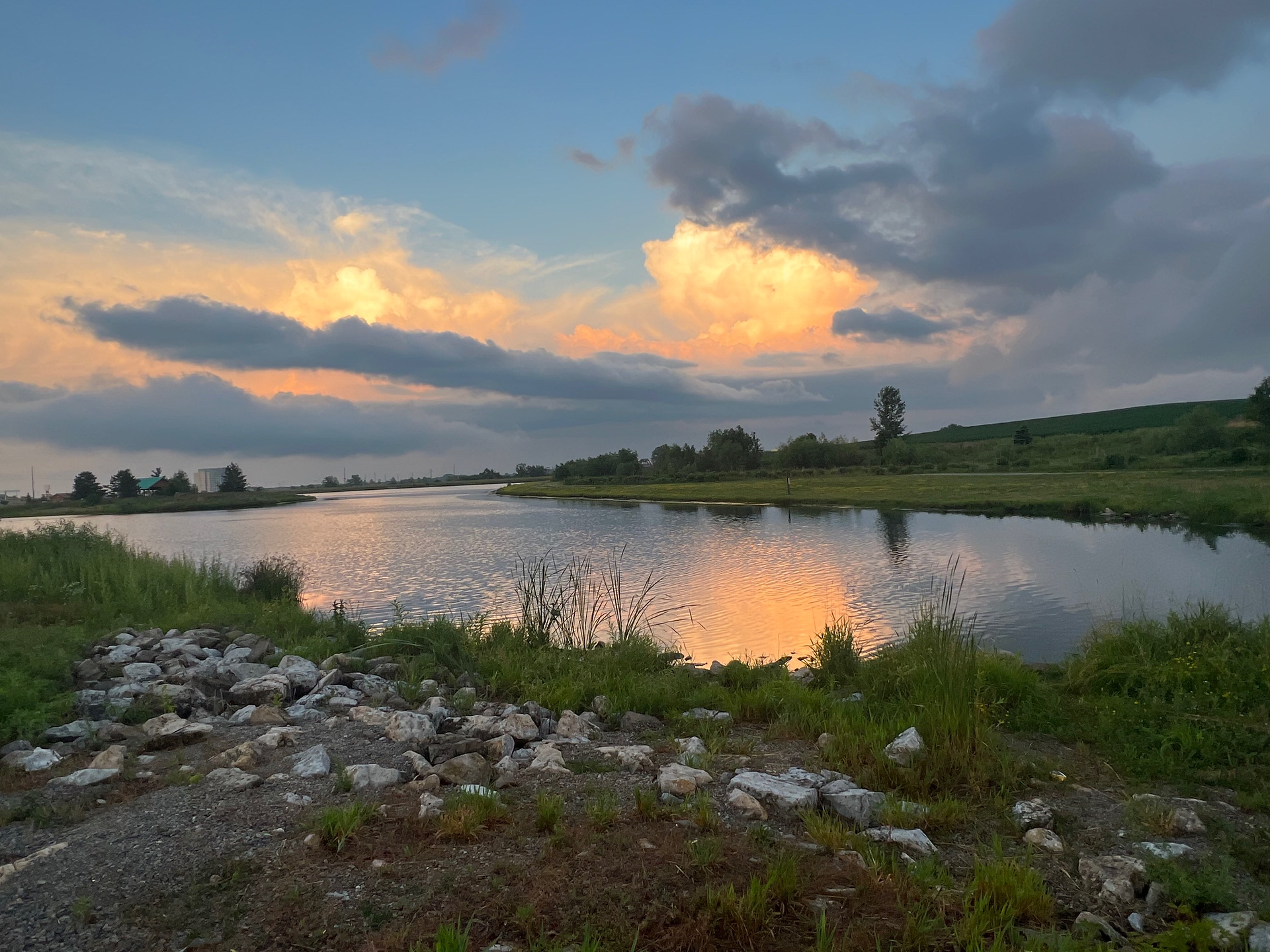 A serene pond with still water reflecting the vibrant colors of the setting sun. The sky is adorned with dramatic clouds illuminated in shades of orange and pink. The foreground features a rocky shore with patches of green grass and reeds, while the background includes a grassy landscape with trees and gentle hills. The peaceful atmosphere is enhanced by the natural beauty and the calmness of the water at dusk.