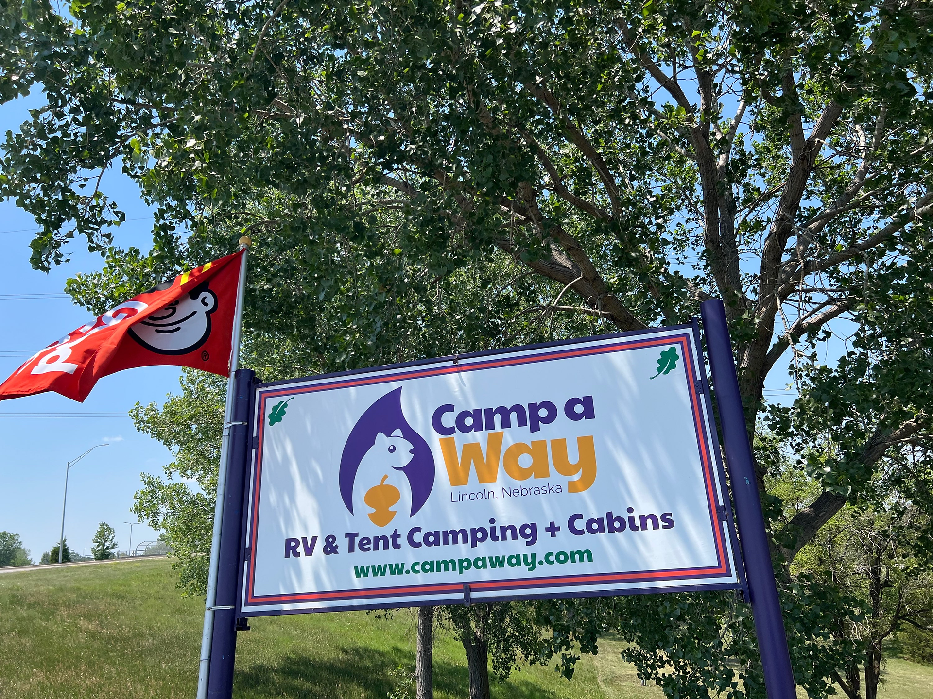 A sign for "Camp a Way" in Lincoln, Nebraska, stands against a backdrop of green trees and a clear blue sky. The sign features a purple and white design with a cartoon chipmunk holding an acorn and includes the text "RV & Tent Camping + Cabins" and the website "www.campaway.com." A red flag with the Good Sam logo, depicting a smiling face with a halo, flutters in the breeze from a pole attached to the sign. The scene conveys a welcoming atmosphere for campers and visitors.