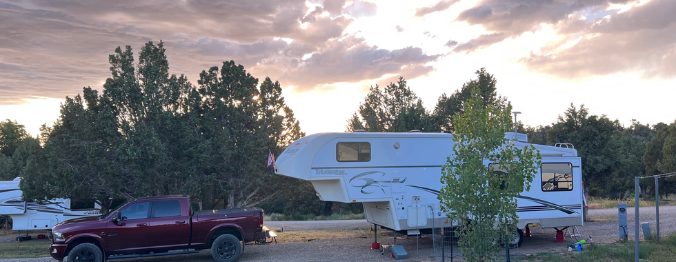 RV campsite at The Views RV Park and Campground featuring a parked white camper and a red truck. The scene is set during sunset with a dramatic sky filled with clouds and golden light filtering through, creating a peaceful and picturesque atmosphere. The campsite is surrounded by trees and greenery.