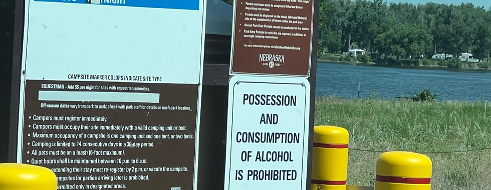 A sign at a campsite registration area displaying the following information: "VISITORS REGISTER HERE. Park Permit AND Appropriate Camping Fee are Required. Nebraska Vehicle Daily Park Permit - $6. Non-Nebraska Vehicle Daily Park Permit - $8. Basic - $15/night." Additional details include campsite rules, such as mandatory registration, maximum occupancy, pet leash requirements, and quiet hours. A separate sign indicates that possession and consumption of alcohol are prohibited. The background shows a lake with trees and parked RVs visible in the distance.
