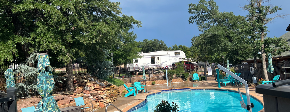 A serene outdoor pool area at a campground, featuring a clear, inviting swimming pool surrounded by turquoise lounge chairs and closed umbrellas with decorative patterns. The pool area is well-landscaped, with trees providing shade and privacy. A small stone waterfall adds a touch of natural beauty to the scene. In the background, a fifth-wheel RV trailer is visible, along with additional camping amenities. The sky is partly cloudy, hinting at a mix of sunshine and possible rain, enhancing the peaceful and relaxing ambiance of the poolside setting.