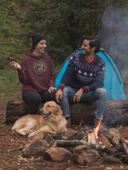 A couple and there dog sitting at a campfire in the woods.