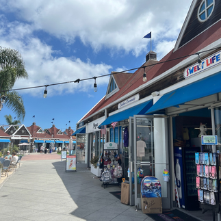Seaside shopping promenade with colorful storefronts and blue awnings, featuring Sweet Life Candies & Gifts, palm trees, outdoor seating, and a sunny coastal atmosphere.