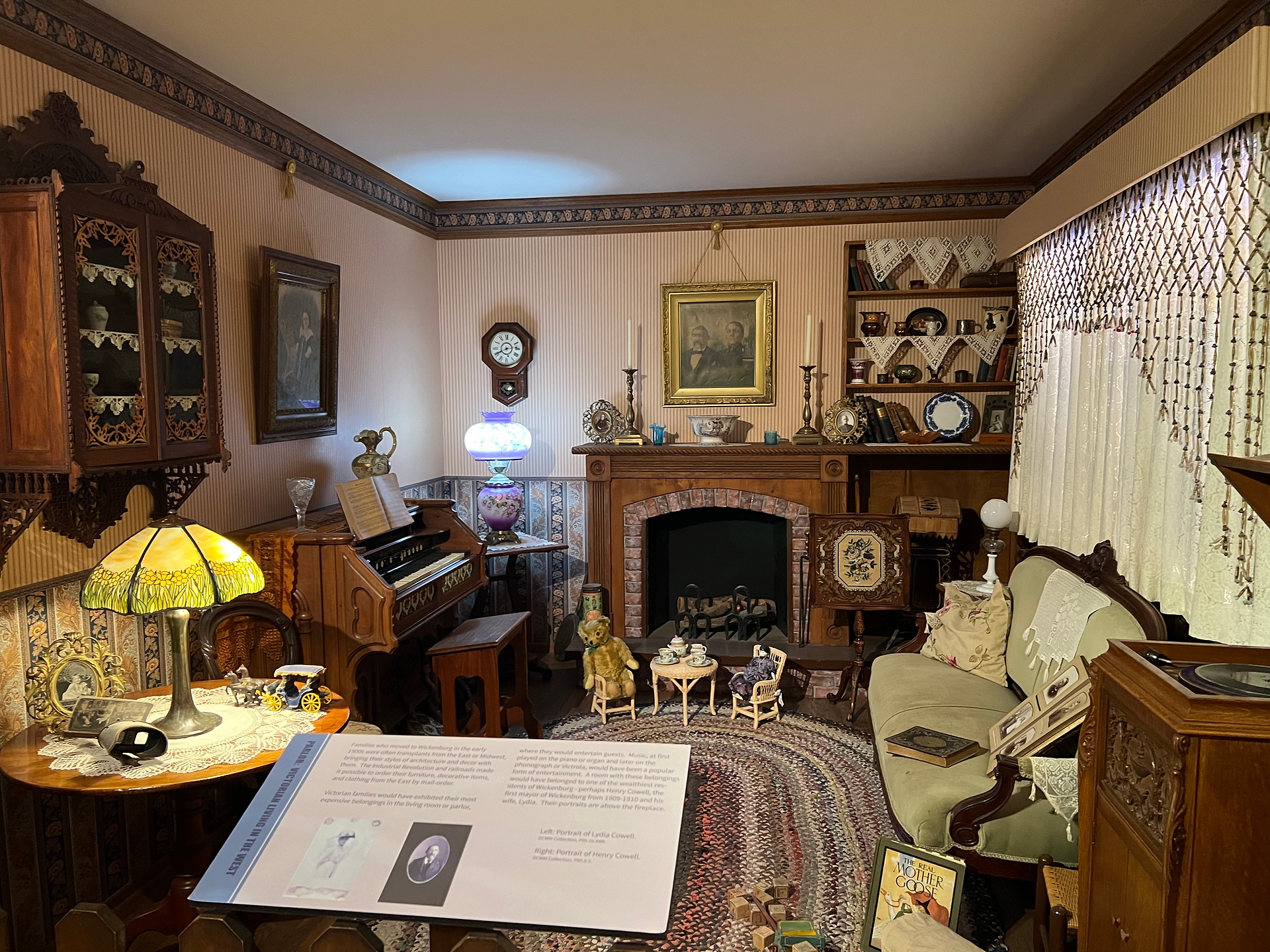 A beautifully restored historical parlor room displayed in a museum. The room features antique furniture, including a sofa, chairs, and a small table set with fine china. A vintage piano and a cabinet filled with glassware are positioned against the walls. The fireplace is adorned with candlesticks, a clock, and framed portraits. The room is illuminated by various antique lamps, and the windows are draped with lace curtains. A display sign provides information about the exhibit.