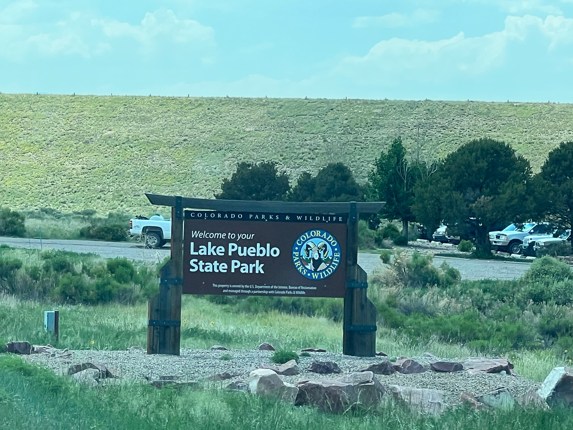 Welcome sign for Lake Pueblo State Park in Colorado Parks & Wildlife, featuring the Colorado Parks & Wildlife logo. The sign is set in a grassy area with a few parked vehicles and trees in the background, under a clear blue sky with scattered clouds.