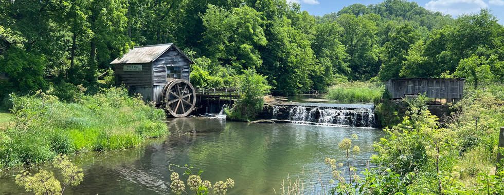 The image shows a picturesque, serene scene featuring an old wooden mill with a large waterwheel, set beside a small waterfall and a calm river. The mill, labeled "Cable Mill" according to a sign on its side, has weathered wooden planks and a shingled roof, giving it a charming, historic appearance.  The waterwheel is positioned at the edge of the mill, partially submerged in the flowing water. The waterfall cascades gently over a stone dam, creating a soothing backdrop of white water.  Surrounding the mill and the water are lush, green vegetation and trees, adding to the tranquil and natural setting. The sky above is bright and clear with a few scattered clouds, enhancing the idyllic and peaceful atmosphere of this rural scene. The overall composition is one of natural beauty and historic charm, evoking a sense of calm and timelessness.
