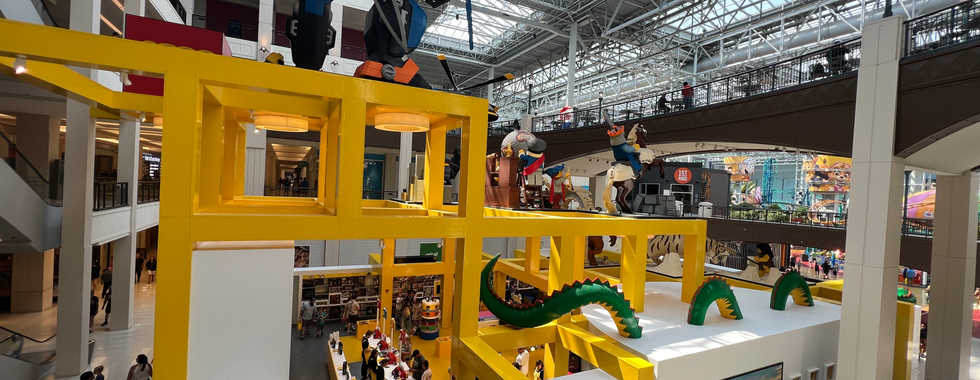 The image showcases a vibrant and busy section of a shopping mall, prominently featuring a large LEGO store. The store has an eye-catching exterior with bright yellow structural elements and massive LEGO sculptures.  On top of the store, there are several large LEGO models, including a giant robot, a pirate with a sword, and other playful characters. These models are incredibly detailed and impressive, demonstrating the creativity and complexity of LEGO constructions. A LEGO dragon is also seen winding its way through the yellow structure, adding an element of fantasy and adventure.  The mall itself has a modern design with high ceilings made of glass, allowing plenty of natural light to flood the space. This creates an open and airy atmosphere. There are multiple levels, with people walking along the corridors and exploring the various shops.  In the background, there is a glimpse of an amusement park or entertainment area within the mall, featuring colorful rides and attractions. This indicates that the mall offers more than just shopping, providing a complete entertainment experience for visitors.  Overall, the image captures a lively and dynamic environment, with the LEGO store serving as a major focal point of interest and excitement within the mall.