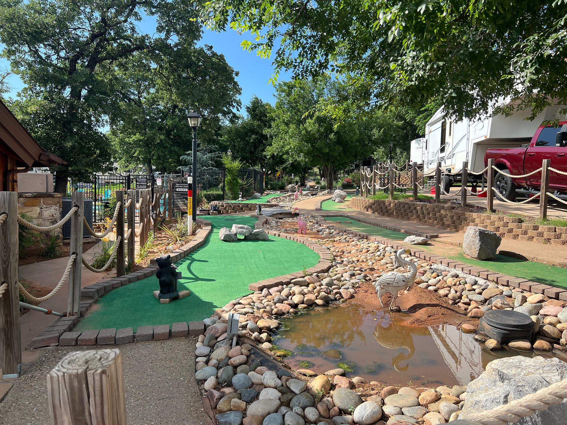 A lively and well-maintained mini-golf course at a campground, featuring pathways bordered by rope fences and various decorations such as a small bear statue and a heron sculpture in a pond. The course includes green artificial turf, rocks, and small bridges, providing a fun and engaging play area. In the background, a red pickup truck hitched to a fifth-wheel RV trailer is visible. The area is shaded by large trees, creating a pleasant and inviting atmosphere under a bright, sunny sky.