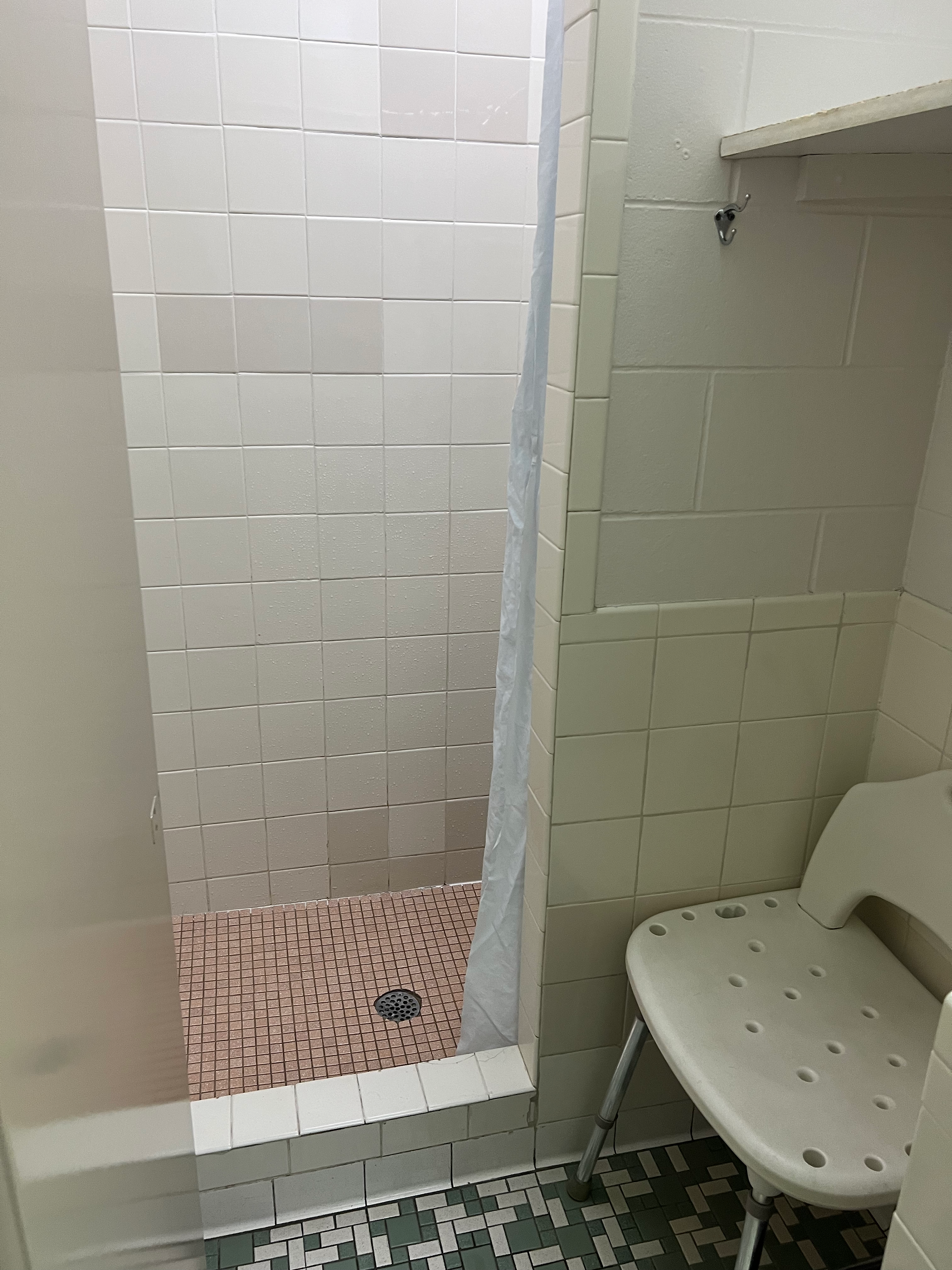 This image shows a clean and well-maintained shower area, likely in a public or shared facility. Here are the key details:  Shower Area: The shower is enclosed with light-colored tiles, and a white shower curtain is partially visible, providing privacy.  Shower Floor: The shower floor has small pinkish tiles with a central drain, ensuring water flows properly.  Adjacent Area: Next to the shower, there is a white plastic shower chair, indicating the facility is accessible and accommodates individuals who may need to sit while showering.  Hooks and Shelves: There are hooks and a small shelf on the wall, offering convenient spots to hang towels and place toiletries.  Flooring: The floor outside the shower has green and white mosaic tiles, adding a decorative touch and likely providing a non-slip surface.  Overall, the image depicts a thoughtfully designed and functional shower space that is accessible and user-friendly.
