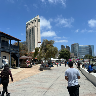Embarcadero walkway with strolling visitors, a historic balconied building, and downtown San Diego high-rises behind.