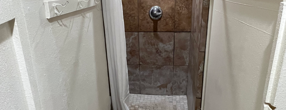 The image shows a small shower stall in a public restroom. The stall has beige and brown tiled walls with a decorative tile border. The showerhead and controls are mounted on the wall in the middle of the stall. A white curtain provides privacy, and there is a small bench just outside the shower area. On the left wall, there are several hooks for hanging towels or clothes. The floor of the shower is also tiled, with a central drain. The overall look is clean and functional.