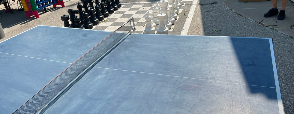 This image showcases an outdoor recreational area equipped with various games, including:  Ping Pong Table: In the foreground, there is a ping pong table set up for outdoor use. It appears to be well-used but in good condition.  Giant Chess Set: In the middle ground, there is a large chess set with oversized black and white pieces arranged on a checkered board painted on the ground, providing a fun and engaging activity for visitors.  Connect Four Game: On the left side of the image, a giant Connect Four game stands, offering another entertaining option for people of all ages.  Seating and Tables: Picnic tables and other seating arrangements are visible, suggesting a comfortable place for people to relax and enjoy the activities.  Other Amenities: The background shows additional amenities and recreational options, indicating a well-equipped and family-friendly outdoor space.  This setup is ideal for encouraging social interaction and outdoor play among visitors, making it a great spot for families and groups.