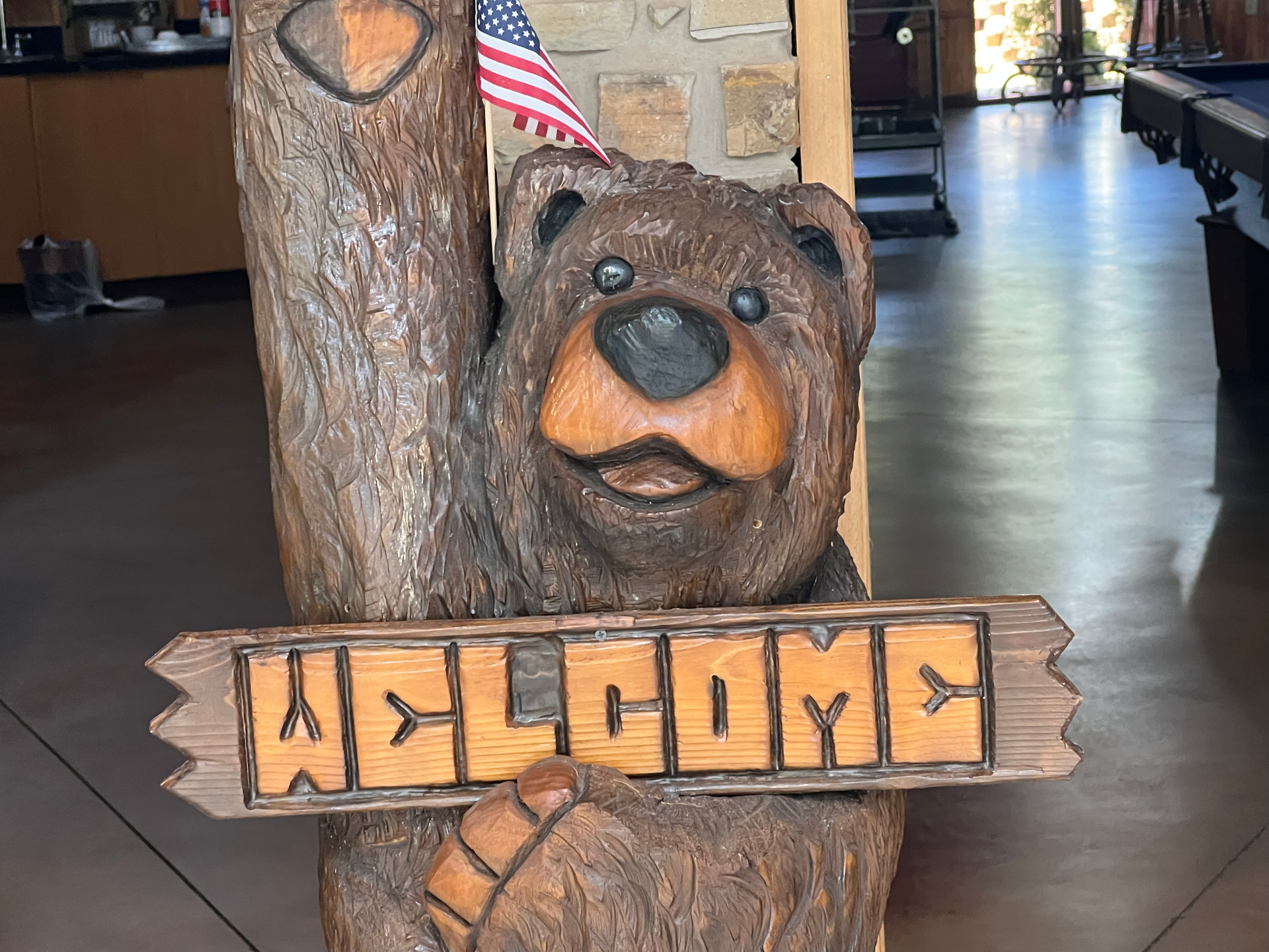 A close-up view of a charming wooden bear carving holding a "Welcome" sign. The bear has a friendly, smiling expression and is also holding a small American flag. The detailed carving showcases the bear's fur and features, adding a rustic and inviting touch to the campground's decor. In the background, a pool table and parts of the recreation room can be seen, enhancing the welcoming atmosphere for guests.