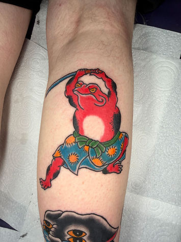 Japanese traditional tattoo on a calf. A red toad in a blue patterned wrap with a green sash, holding a sword above its head.