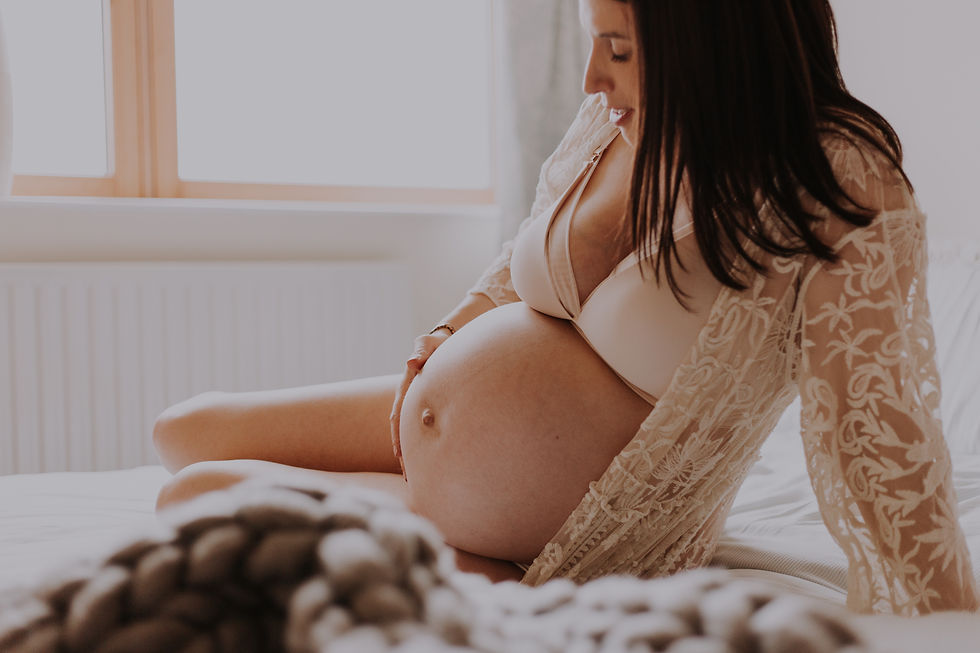 Pregnant woman cradling her baby bump during a maternity photoshoot, wearing a white bra and lace robe in soft natural light.