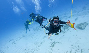Un instructor de buceo guía a los estudiantes en una inmersión a la deriva, sosteniendo un DSMB en un entorno de agua azul clara.