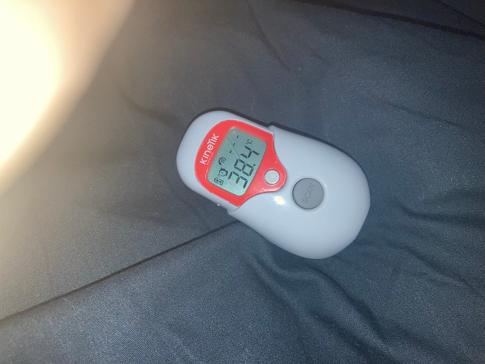 An electronic thermometer in white and red plastic with grey buttons. It has the brand name "Kinetix" written above a digital readout screen. The screen reads "38.4 c"